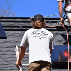 A solar installer from Southern Energy Management climbs a ladder onto a roof where solar panels are being installed. He wears a shirt that reads "Southern Energy Solar Crew – Lead the Change," and a black mesh cap. Another worker is visible adjusting equipment on the roof under a clear blue sky.