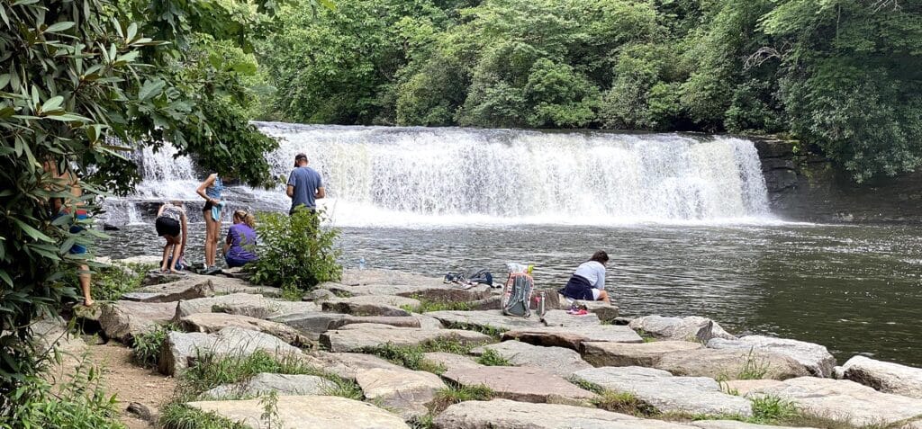 People relax and enjoy a day out near Hooker Falls, a wide and picturesque waterfall that spans across a river, surrounded by large rocks and lush trees