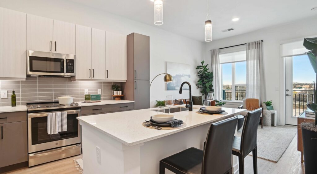 A modern kitchen with a central island, bar stools, pendant lighting, and stainless steel appliances. The room is bright with natural light coming from a window and glass door leading to a balcony.