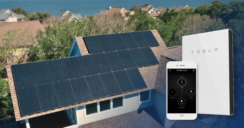 Rooftop solar panel installation on a house near the coast with a Tesla Powerwall and a smartphone displaying the Tesla Power Flow app.