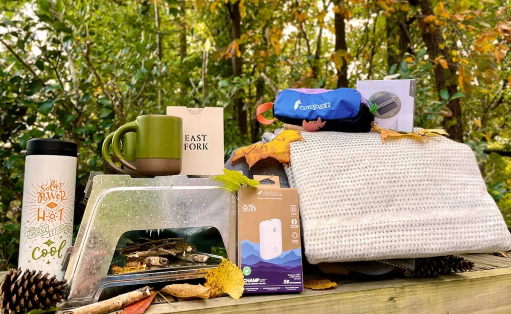 Selection of B Corp items including a kanteen, mug, blanket, portable fire pit, portable charger, and dopp kit with fall trees in the background