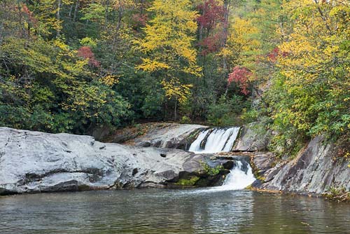 Hunt Fish Falls gently flows over a smooth rock face into a calm river, framed by vibrant autumn foliage, showcasing the serene beauty of the season