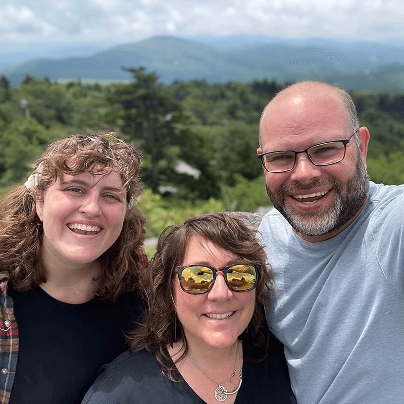 Laurie smiling with her family during an outdoor adventure with green trees and mountains in the distance