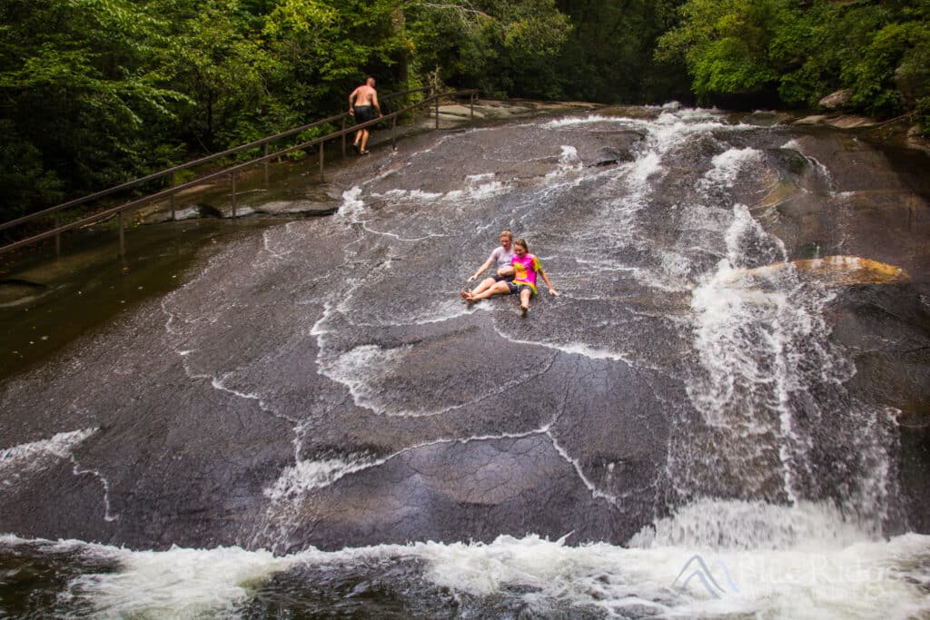 Two children enjoy the thrill of sliding down Sliding Rock, a natural water slide formed by a wide, smooth rock in a lush, verdant landscape.