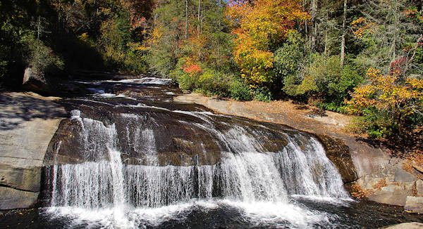 Turtleback Falls gently flows across a broad, flat rock surface surrounded by dense, colorful autumn trees, creating a picturesque natural cascade